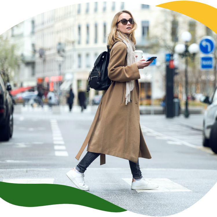 Moderní žena s velkými slunečními brýlemi a v dlouhém béžovém plášti přechází přes ulici a v pravé ruce drží mobilní telefon.
