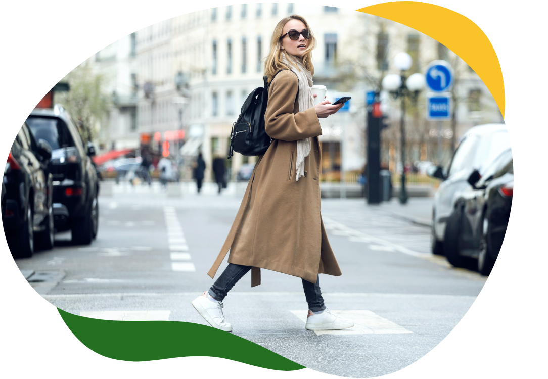 Moderní žena s velkými slunečními brýlemi a v dlouhém béžovém plášti přechází přes ulici a v pravé ruce drží mobilní telefon.