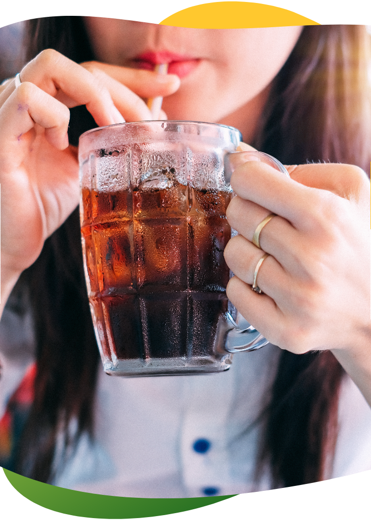 Mladá žena s dlouhými hnědými vlasy drží v ruce vysokou sklenici a brčkem z ní popíjí sycený nápoj.