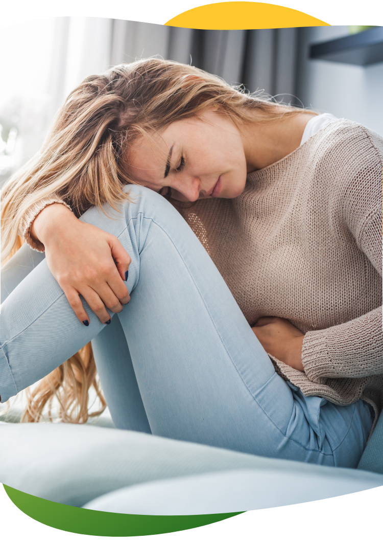Mladá žena, kterou trápí nadýmání během menstruace, sedí skrčená na zemi a levou rukou se drží za břicho.